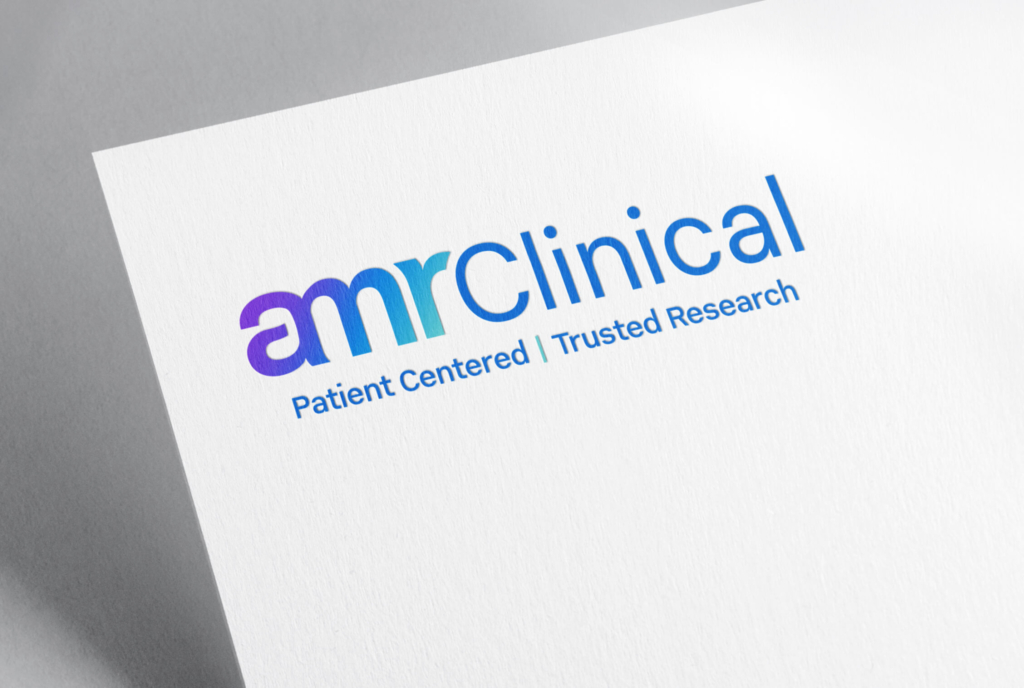 A logo mockup on textured white paper featuring the text "amrClinical" in a modern, blue-to-purple gradient font for "amr" and solid blue for "Clinical." Below the logo, the tagline reads "Patient Centered | Trusted Research" in blue text, separated by a thin vertical teal line. The design conveys a clean and professional look suitable for a clinical or healthcare research brand.