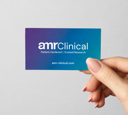 A hand holding a business card featuring the "amrClinical" logo in white on a gradient background transitioning from purple to teal. Below the logo, the tagline reads "Patient Centered | Trusted Research," with the website "amr-clinical.com" centered at the bottom.