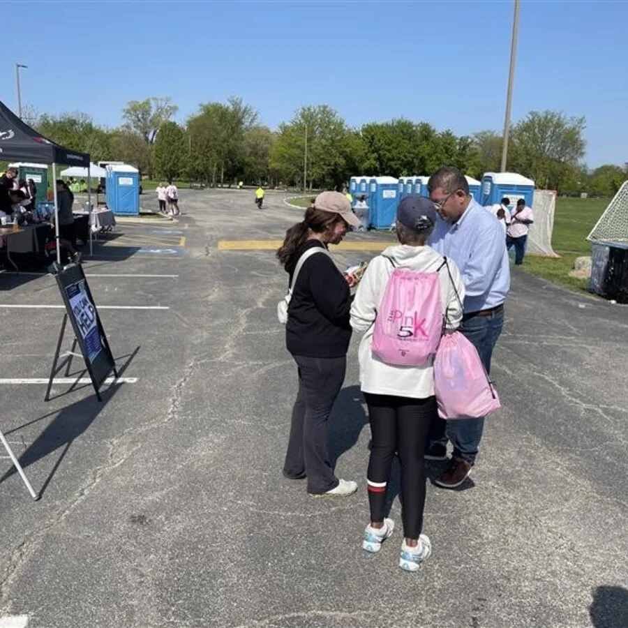 Three people are seen talking in a parking lot near the event area. Two women wearing caps and casual clothing, one with a pink “Pink 5K” drawstring bag, are speaking with a man in a light blue shirt. In the background are portable toilets, more attendees, and informational tents under a clear blue sky.