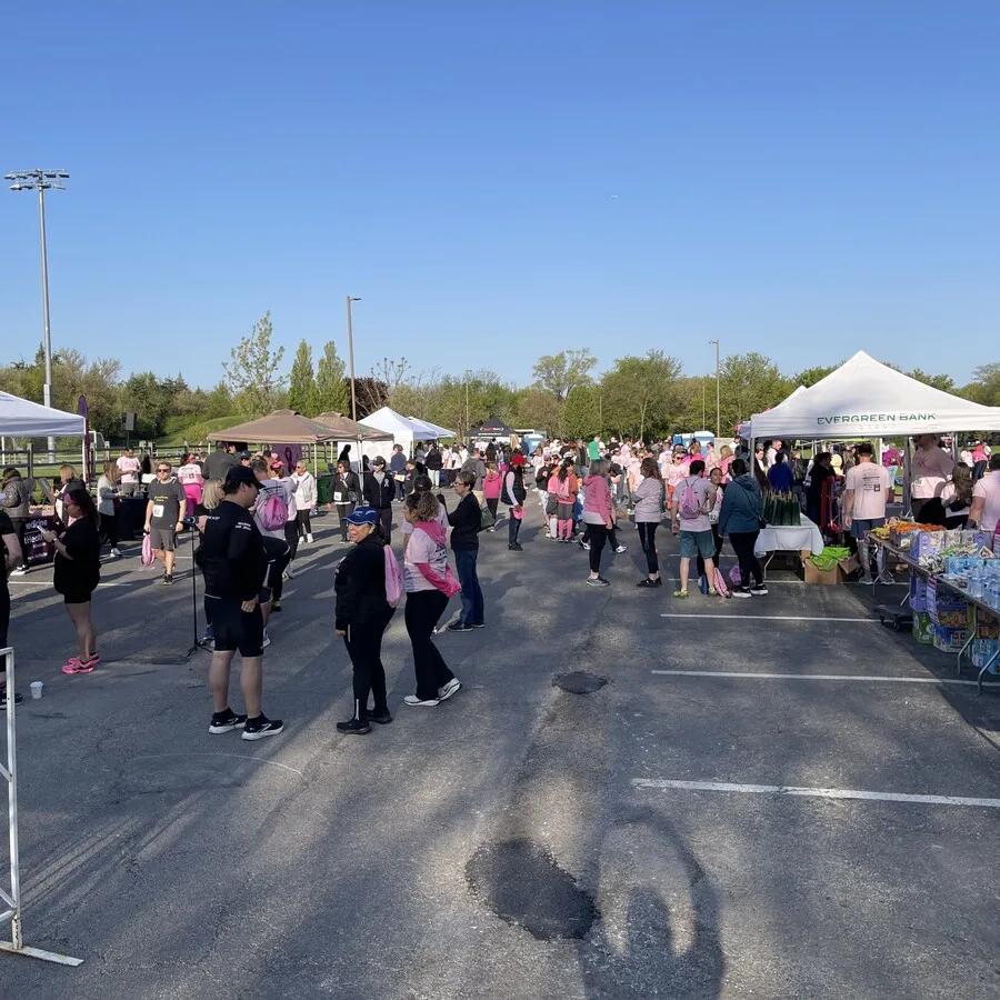 Wide shot of a busy parking lot at the Pink 5K event, filled with attendees wearing pink shirts and gear. Tents and booths line both sides of the area, including one from Evergreen Bank. People are gathered, mingling, and preparing for or finishing the event. The mood is lively and festive.