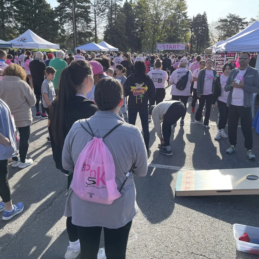 Crowd gathered near the “START” line of the Pink 5K event, with many participants wearing pink t-shirts and drawstring backpacks. In the foreground, a person with a pink “Pink 5K” backpack stands next to a cornhole game board. The path is surrounded by vendor tents and the scene is bright with morning sunlight.