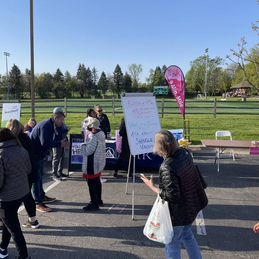 Busy scene at the AMR booth during the Pink 5K event. A whiteboard in the foreground promotes a raffle and shingles vaccine, while people visit the booth to talk and gather information. AMR staff are behind the table under a clear blue sky, and a green athletic field with trees runs along the back of the display area.