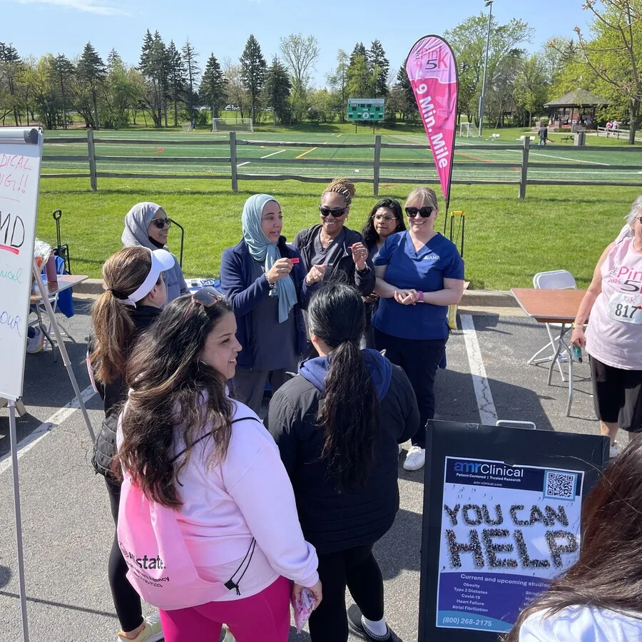 A group of women, including AMR staff and attendees, are gathered in conversation near the AMR booth at the Pink 5K. A “9 Min Mile” feather flag and a green field are in the background. One woman in a light blue headscarf appears to be giving information or a demonstration. A sandwich board with “YOU CAN HELP” and clinical trial details is partially visible on the right.