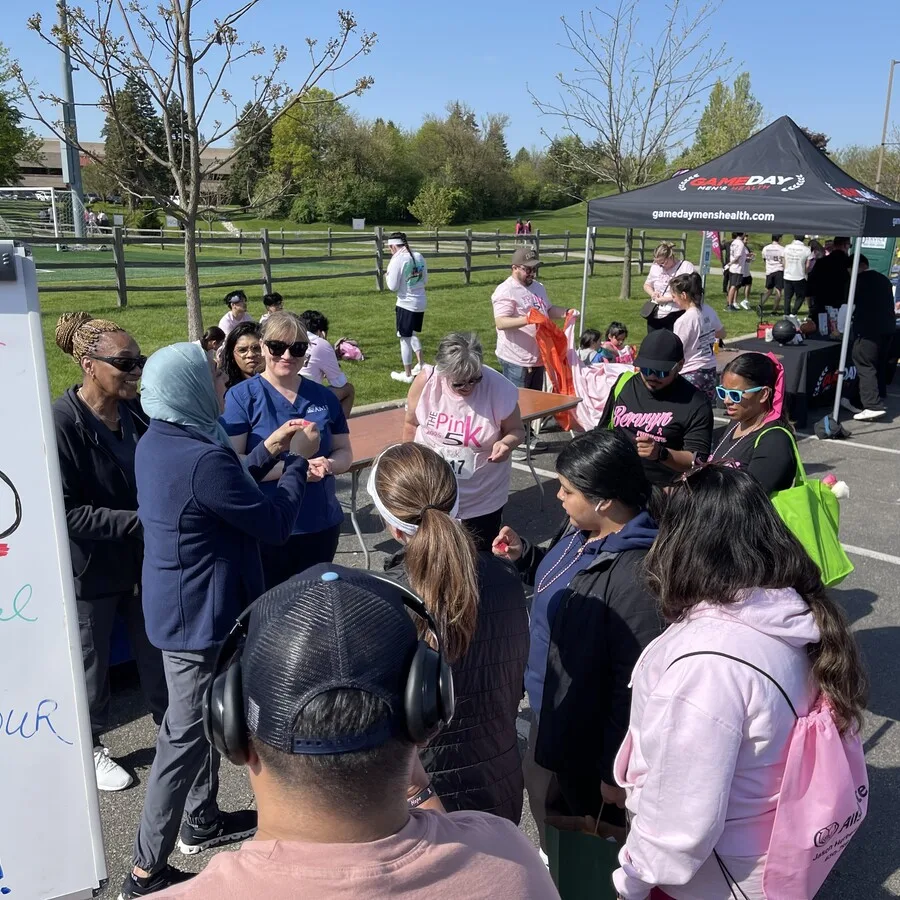Event attendees gather closely around the AMR booth, listening to a woman in a headscarf who is speaking and holding a small item. AMR team members in branded clothing stand nearby. Many attendees wear Pink 5K t-shirts and carry drawstring bags. A GameDay Men’s Health tent is visible in the background.