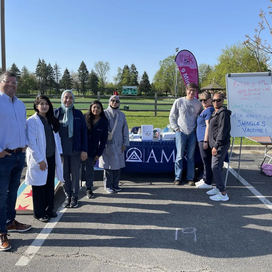 Group photo of eight individuals standing in front of an AMR informational table set up outdoors in a parking lot. A blue tablecloth with the AMR logo covers the table, which includes flyers and health materials. A sign to the right promotes a raffle and shingles vaccine, and a pink “Pink 5K” feather flag is visible. The background shows a green field and trees.