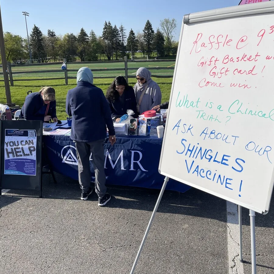 Close-up of the AMR booth at the Pink 5K event. Several team members are behind the table, interacting with visitors. The table has pamphlets, giveaway items, and a display board reading “YOU CAN HELP” from amrClinical. In the foreground, a whiteboard promotes a raffle, clinical trials, and shingles vaccines in colorful writing. The event takes place beside a field lined with trees and a fence.