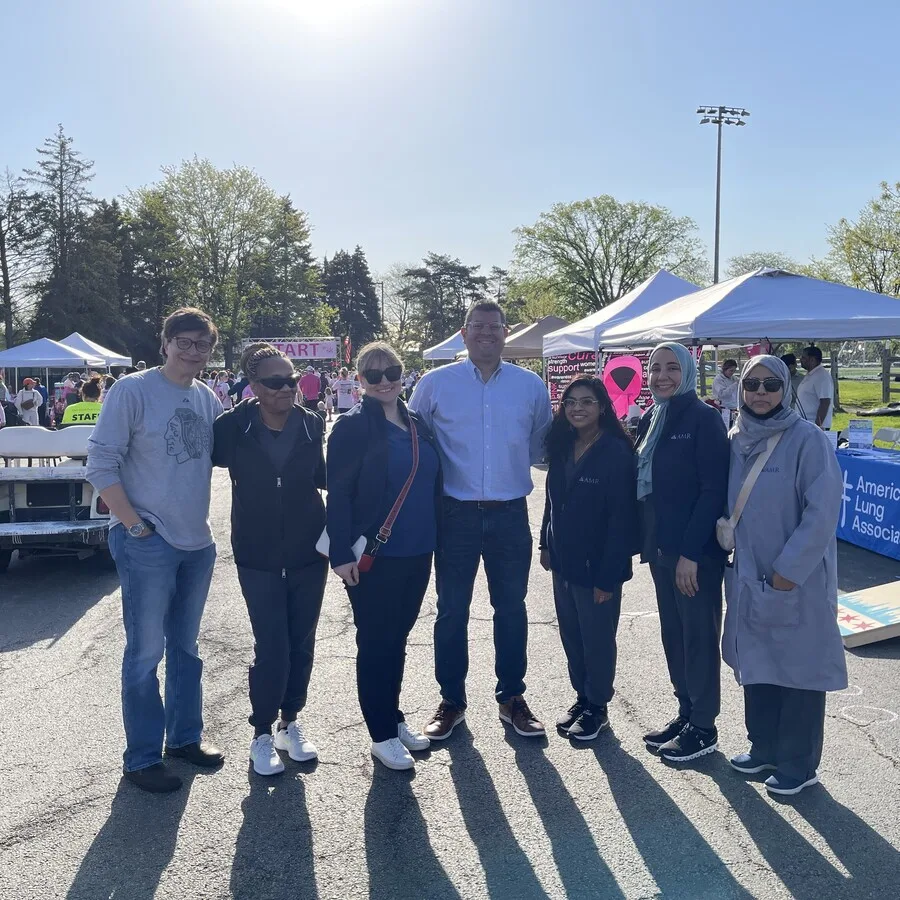 A group of seven people pose in front of tents and informational booths at the Pink 5K event. They are smiling, standing in front of the “START” banner in the background, surrounded by tents including one from the American Lung Association. The group includes AMR team members and other participants under clear blue skies.