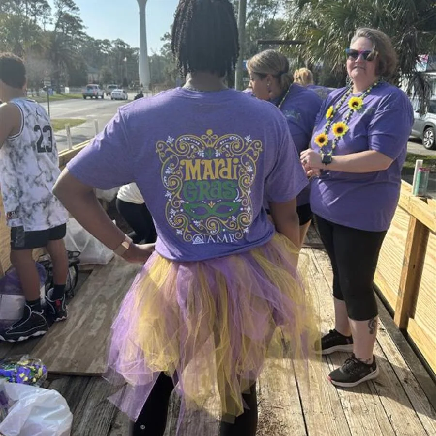A group of people stands on a wooden parade float deck preparing for a Mardi Gras event. The focus is on a woman wearing a purple AMR Mardi Gras t-shirt with decorative lettering and a yellow and purple tulle tutu. Another woman in sunglasses and a matching AMR shirt adorned with sunflower necklaces is talking nearby. The background includes palm trees, a water tower, and others in festive attire.