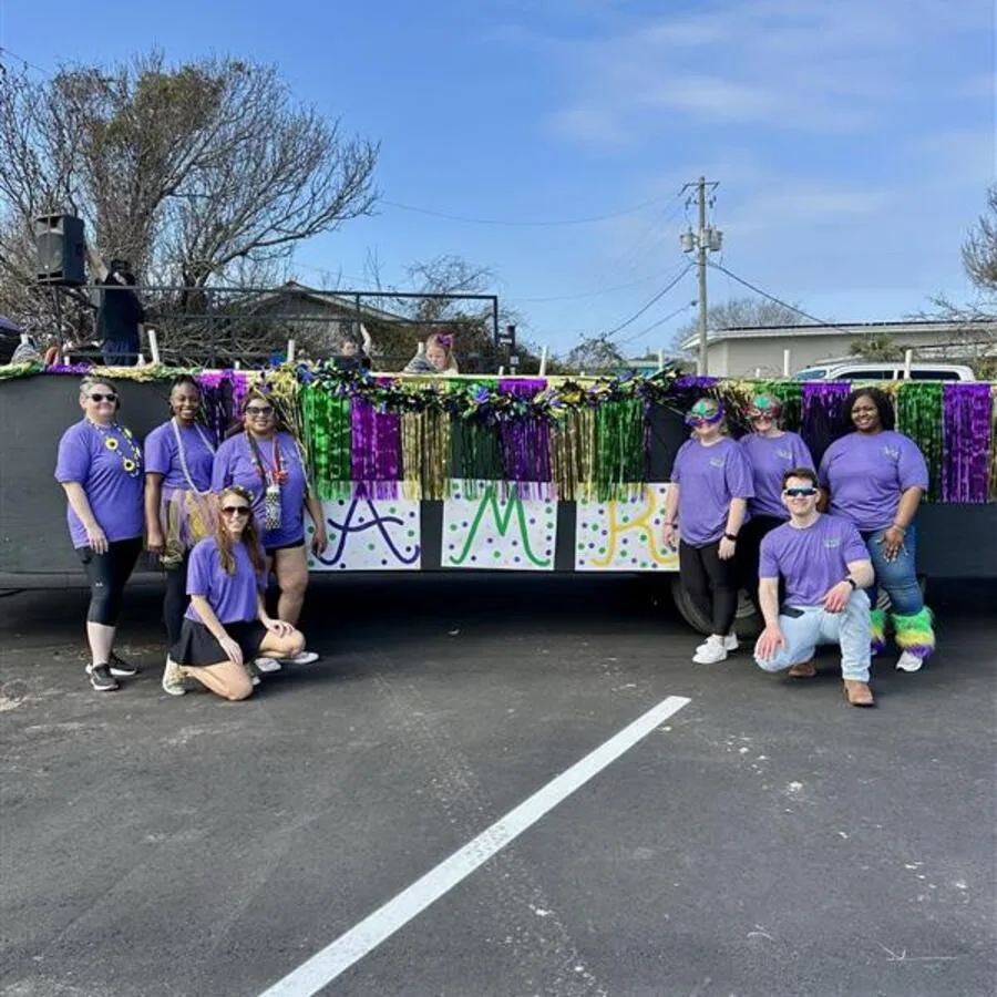 A group of AMR team members in matching purple Mardi Gras t-shirts pose in front of a decorated parade float. The float is draped with metallic green, gold, and purple fringe, and signs spelling out “AMR” are attached to the side. Some participants wear beads, fun sunglasses, and colorful accessories, standing or kneeling on an asphalt parking lot under a bright blue sky.