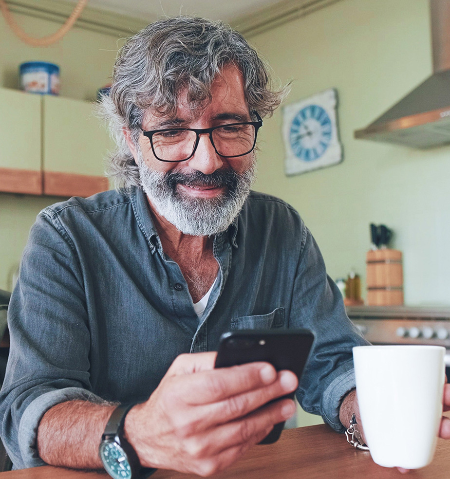 A middle-aged man with salt-and-pepper hair, beard, and black-frame glasses sits at a wooden kitchen table, holding a smartphone in one hand and a white coffee mug in the other, smiling as he reads from his phone.
