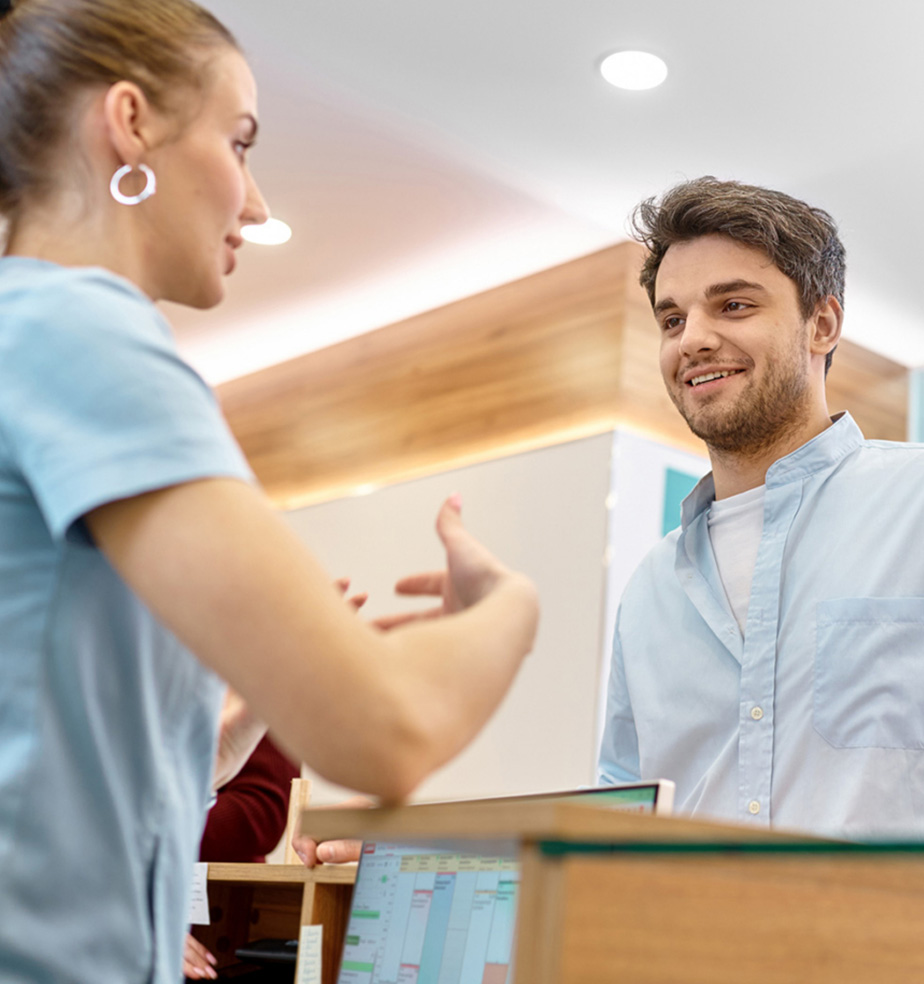 A friendly interaction at a clinic reception desk: a female medical receptionist or nurse (in light blue scrubs) gestures with her hands while speaking to a smiling male patient standing on the other side, with a computer monitor and appointment calendar visible in the foreground.