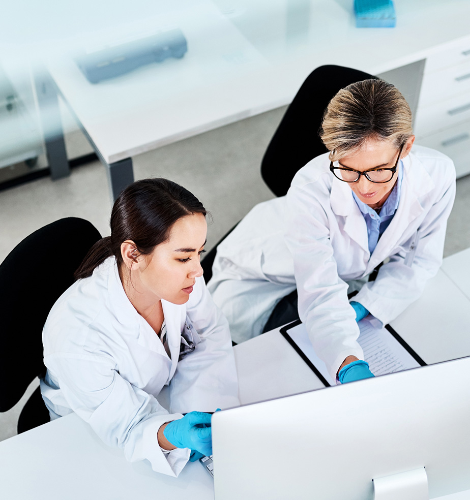 Two lab professionals—one younger Asian woman and one older Caucasian woman—both in white coats and blue gloves, seated at a desk, leaning in and pointing at a large desktop monitor as they discuss their data.