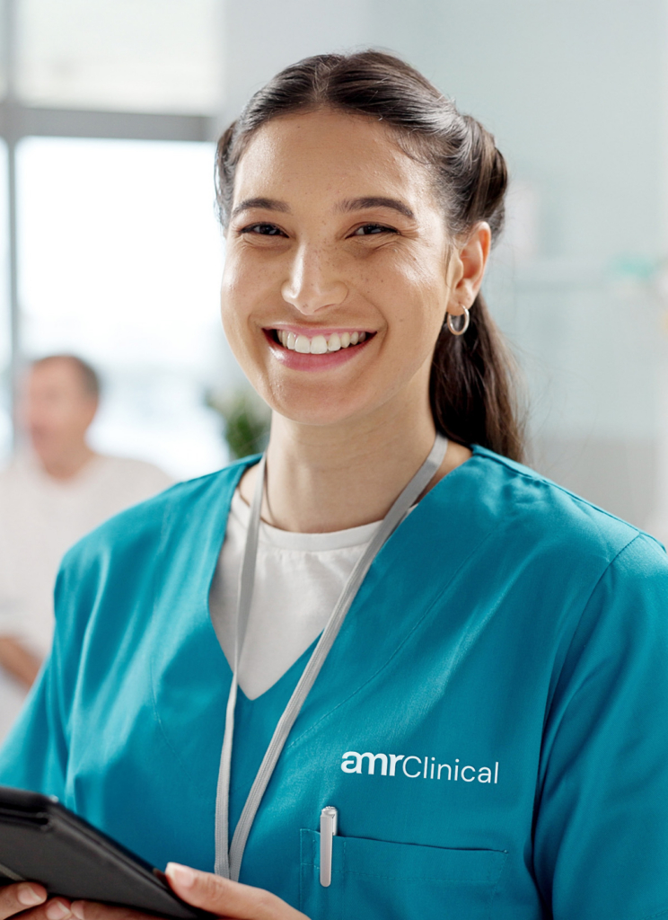 A close-up portrait of a smiling young female healthcare professional with dark hair pulled back, wearing teal “amrClinical” scrubs, a lanyard, and holding a tablet against a softly blurred clinic background.