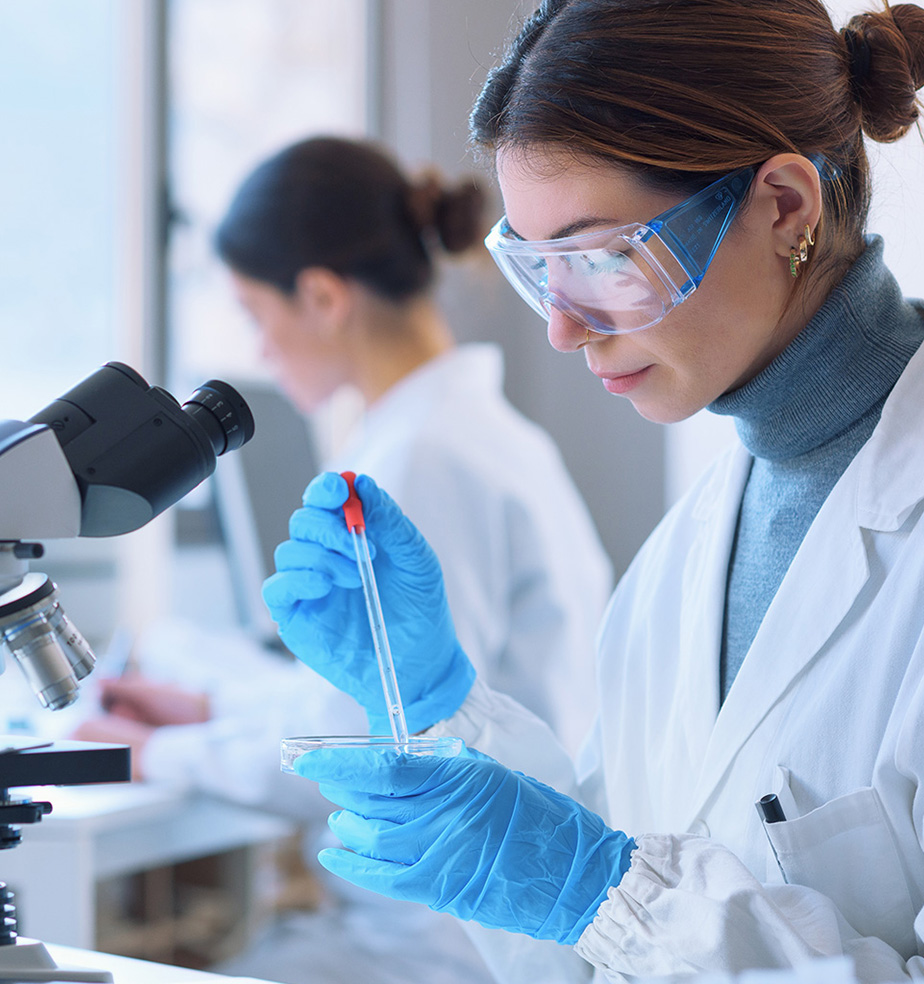 A close-up of a young woman scientist in a white lab coat and blue gloves, wearing clear safety goggles, using a pipette to transfer liquid into a petri dish at a microscope station, with another researcher blurred in the background.