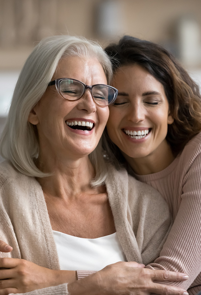 Two women share a warm embrace and laugh together: an older woman with silver hair and glasses wearing a light cardigan, and a younger brunette in a pink sweater wrapped around her from behind, both with their eyes closed and big smiles on their faces.