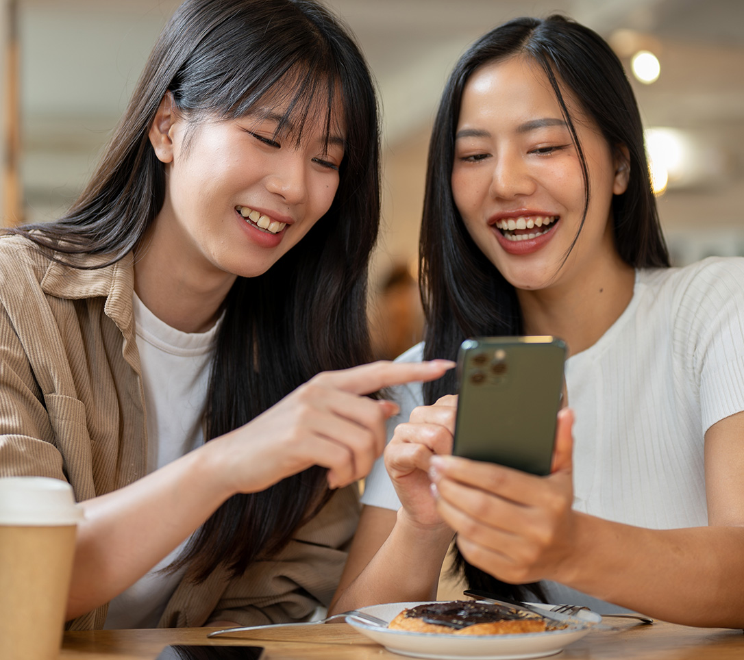 Two young women sitting side by side at a café table, one holding a smartphone while the other points at its screen; both are smiling and looking at the phone, with a coffee cup and a chocolate-topped pastry on the table in front of them.
