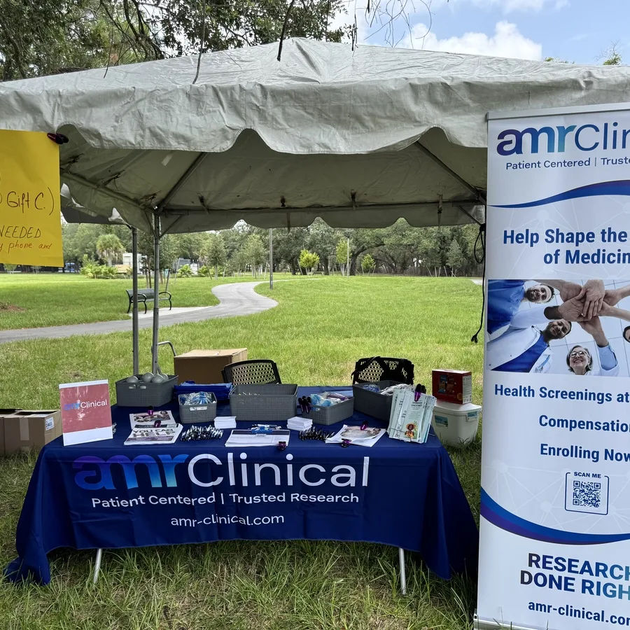 Outdoor informational booth with a navy blue tablecloth labeled “amrClinical - Patient Centered | Trusted Research.” The table is under a canopy and displays promotional materials, pens, brochures, and giveaway baskets. A yellow raffle sign reads “RAFFLE WIN $50 (1 of 2 $50 Gift C.), No purchase needed.” A large vertical banner on the right promotes health screenings at no cost with compensation, stating “Help Shape the Future of Medicine.” The booth is set in a grassy park with a paved path and trees in the background.