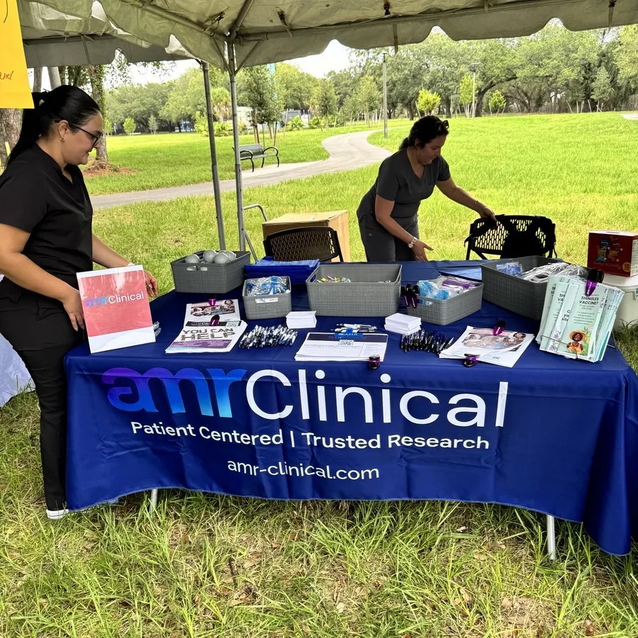 Two women are setting up or organizing items on a table at the amrClinical booth under a canopy in a park. The table is covered in a branded tablecloth and filled with baskets of giveaways, pens, flyers, and a small sign displaying the amrClinical logo. A yellow raffle sign is partially visible to the left. The surrounding area is grassy with trees and a walking path.