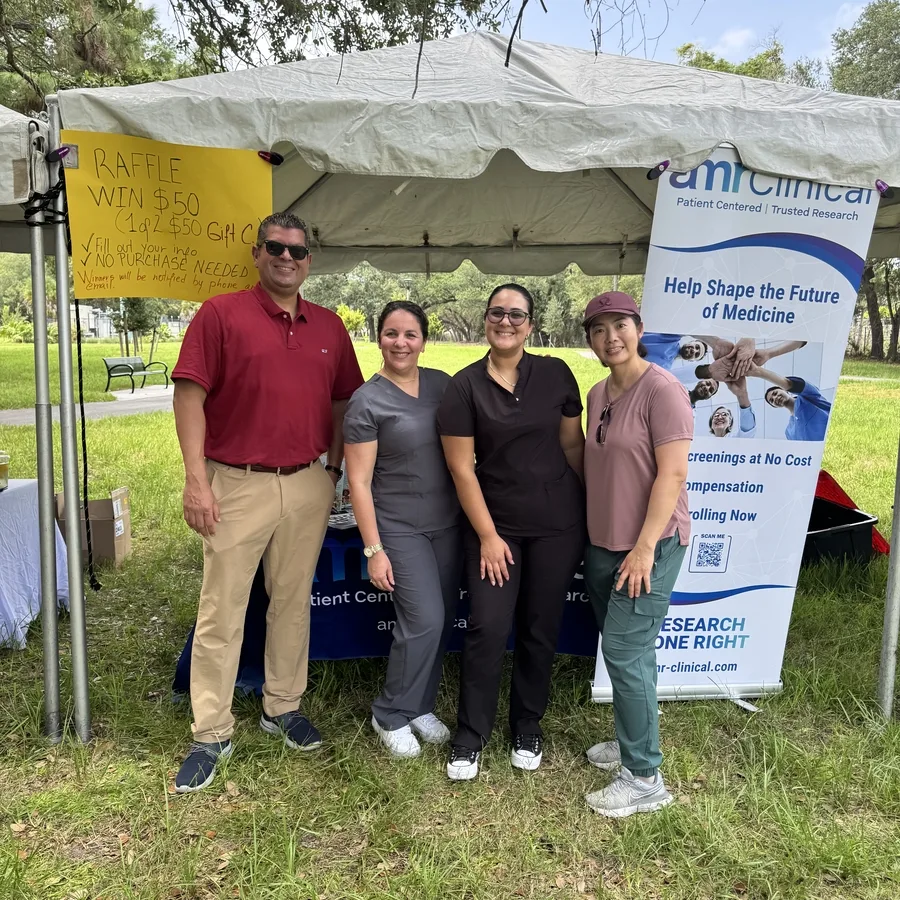 Five people (three women in scrubs, one man in a red polo, and one woman in casual attire) are smiling and standing in front of the amrClinical booth outdoors. The tent has a yellow raffle sign and a tall banner advertising free health screenings and clinical research enrollment. The setting is a grassy park with trees and a walking trail in the background.