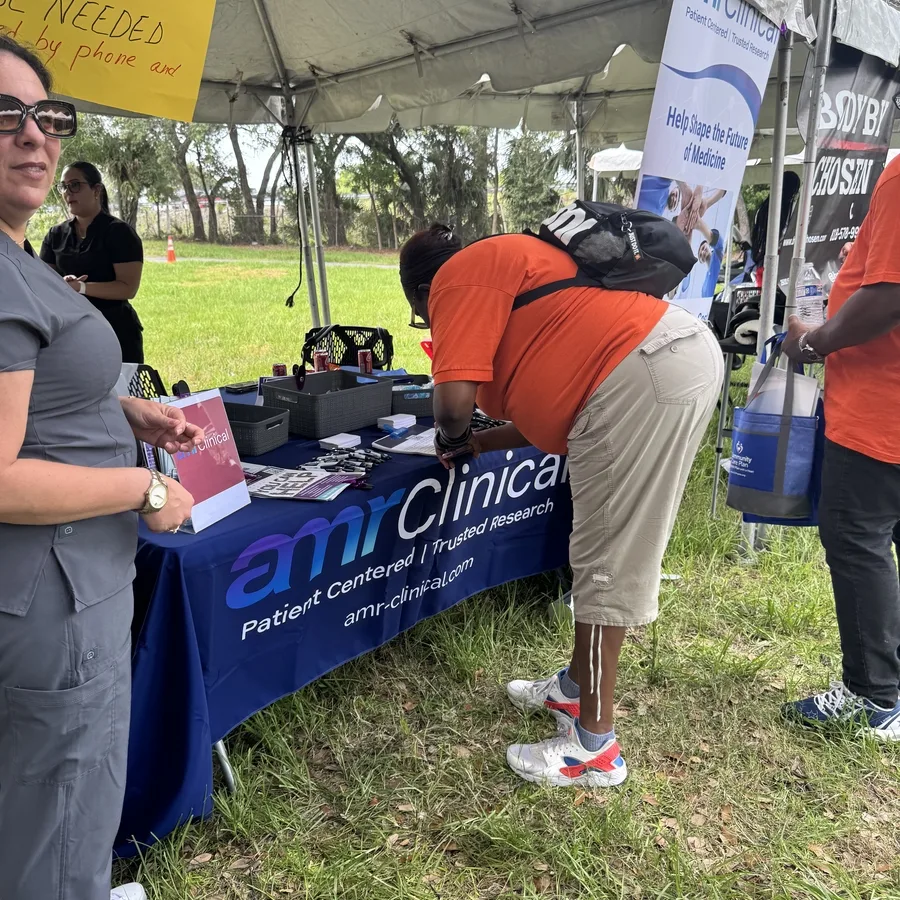 Two women are setting up or organizing items on a table at the amrClinical booth under a canopy in a park. The table is covered in a branded tablecloth and filled with baskets of giveaways, pens, flyers, and a small sign displaying the amrClinical logo. A yellow raffle sign is partially visible to the left. The surrounding area is grassy with trees and a walking path.
