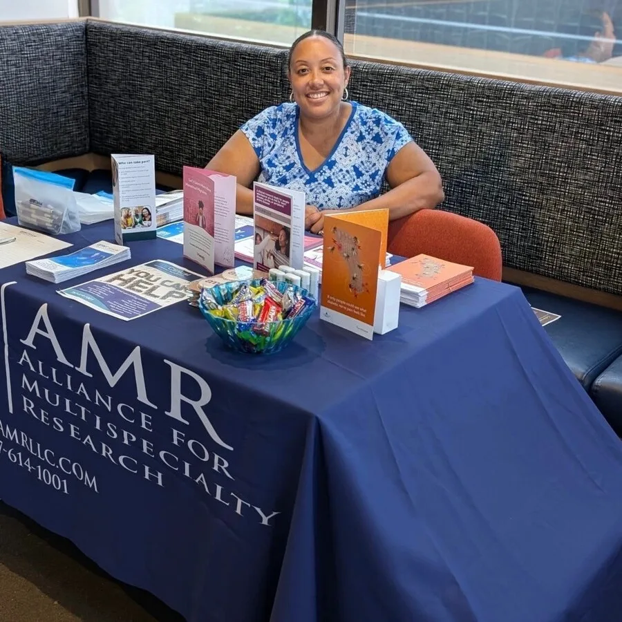 A woman is smiling and seated behind an informational table covered with a navy blue tablecloth that reads “AMR Alliance for Multispecialty Research” along with contact information. The table is indoors and neatly arranged with pamphlets, flyers, a bowl of assorted candy, branded items, and health-related literature. The setting appears to be a lounge or waiting area with cushioned bench seating and natural light from nearby windows.