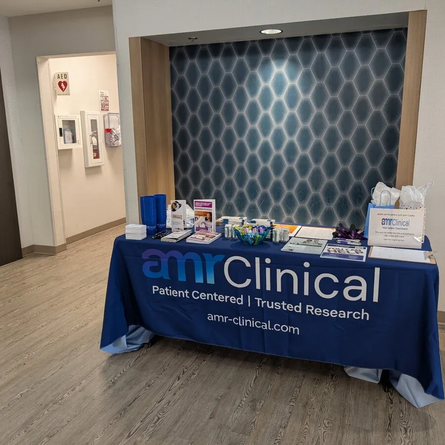 An unattended amrClinical display table set up indoors against a modern wall with a geometric hexagonal pattern. The table is covered with a branded navy blue tablecloth that reads “amrClinical - Patient Centered | Trusted Research.” It is stocked with giveaways including blue cups, pamphlets, pens, candy, and a raffle box labeled “Build with us! Win a gift card.” A hallway with medical emergency equipment is visible to the left.