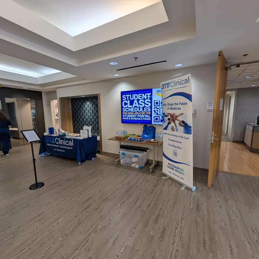 Wide view of an indoor informational setup in a building with wood flooring and modern lighting. On the left, the amrClinical table is positioned near a hallway, displaying branded materials and promotional items. To the right, a tall vertical banner stands next to a table with snacks and water bottles, promoting clinical research participation. Behind the table, a screen displays “Student Class Schedules” and a QR code, suggesting the event is hosted at a student or educational facility.