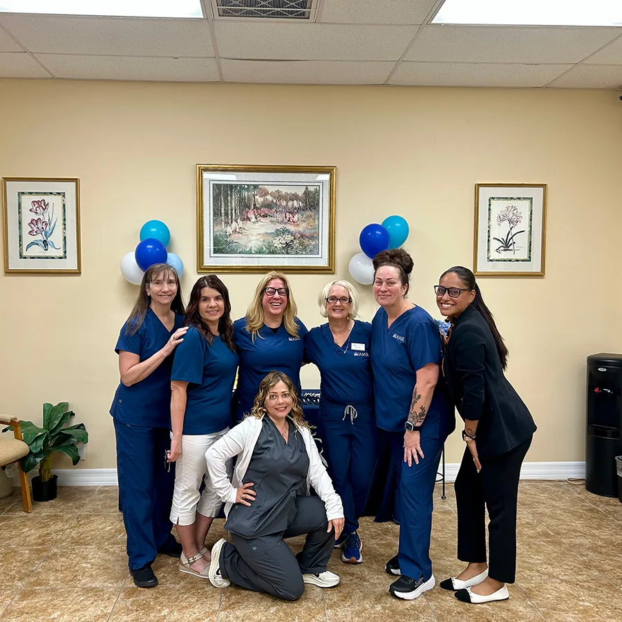 “Seven team members—mostly in navy scrubs, with one in a blazer—smile together in front of the AMR table at an indoor event.”