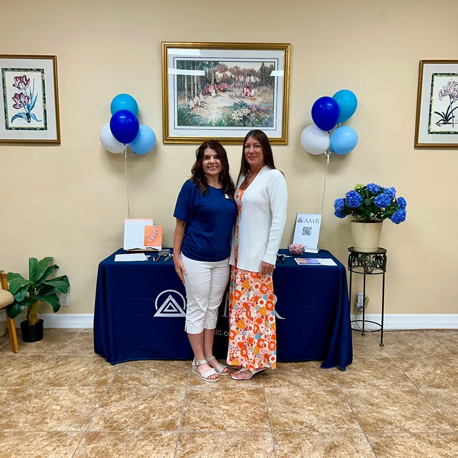 “Two women—one in a navy top and white pants, one in an orange floral dress with a white cardigan—pose in front of the AMR info table with balloons.”