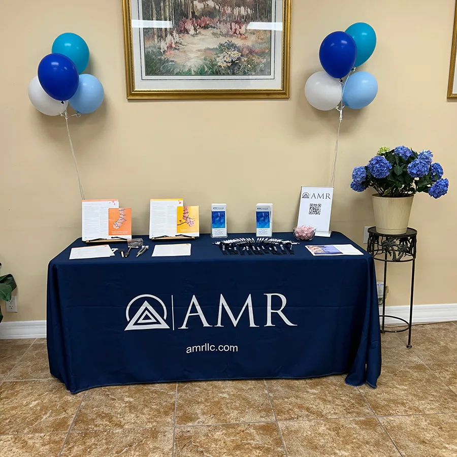 AMR-branded information table in a clinic lobby with brochures, pens, a QR-code sign, blue/white balloons, and a potted blue hydrangea.”