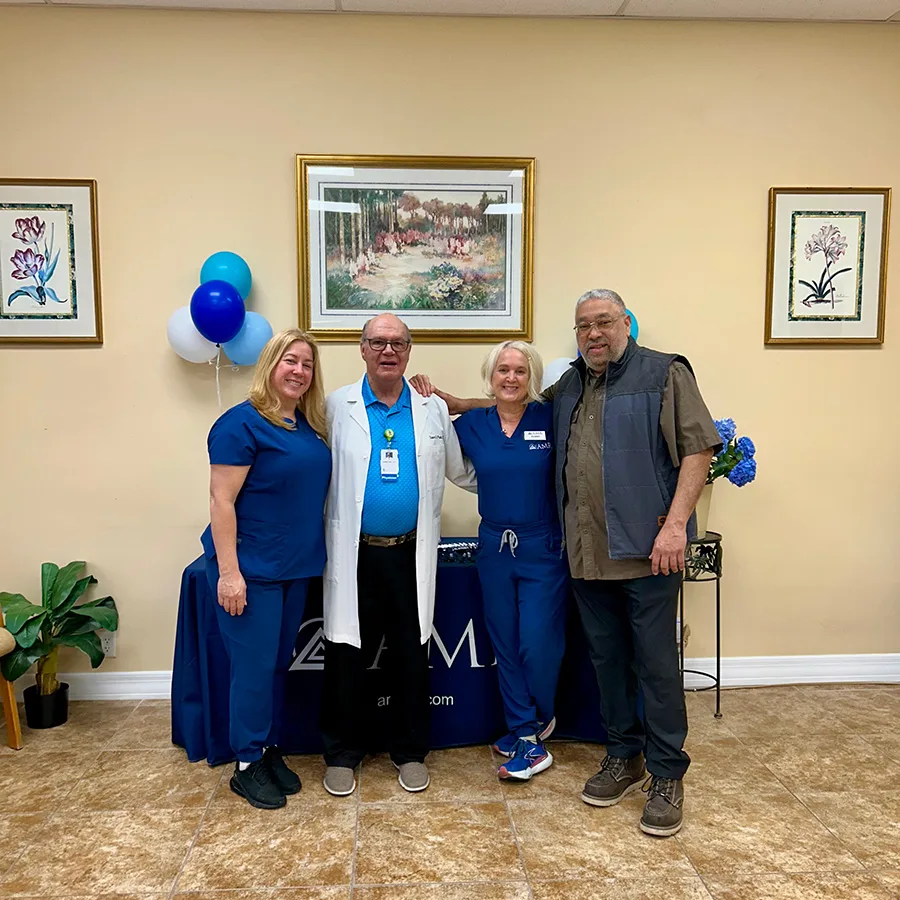 “Four people—two in blue scrubs, a doctor in a white coat, and a guest—standing together in front of the AMR table.”