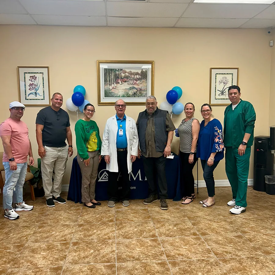 “Group of nine attendees, including a doctor in a white coat, lined up in front of the AMR table decorated with blue and white balloons.”