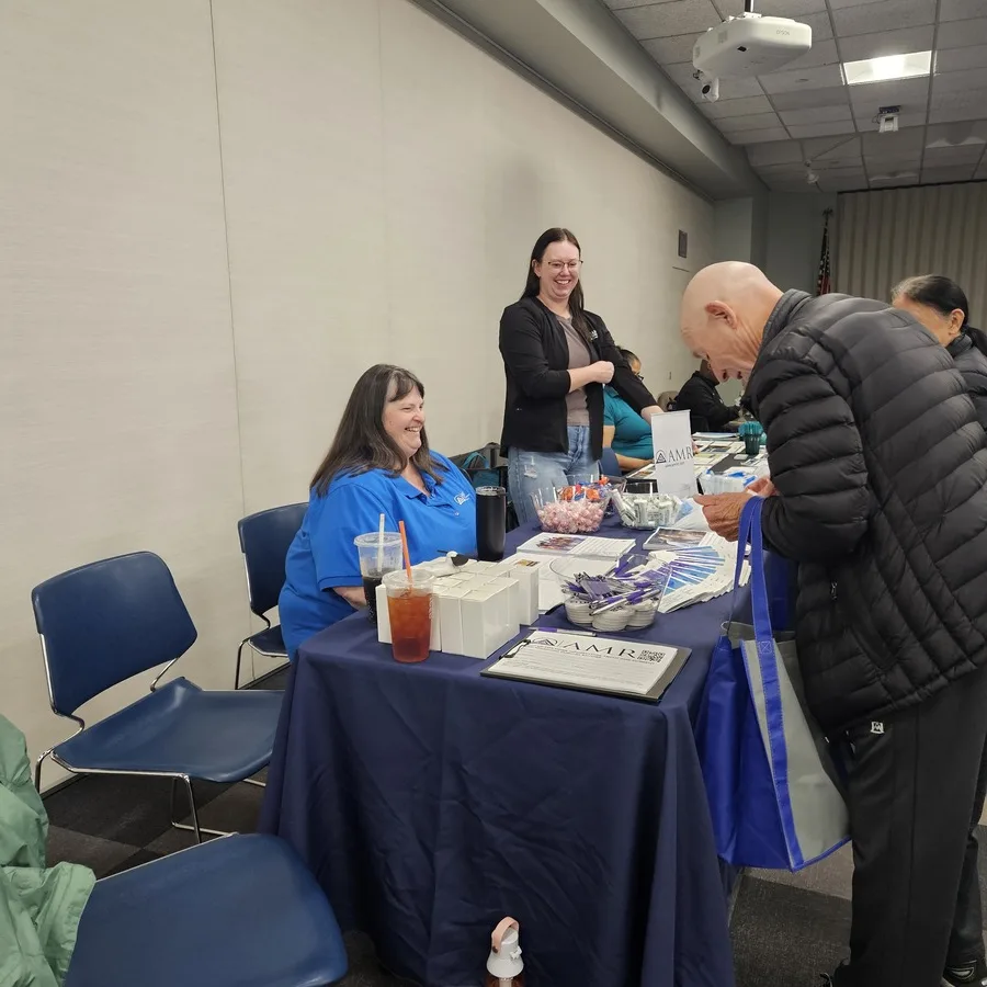 Two AMR representatives, one seated and one standing, interact with an older man at their event booth. Promotional items and brochures are displayed on the table.
