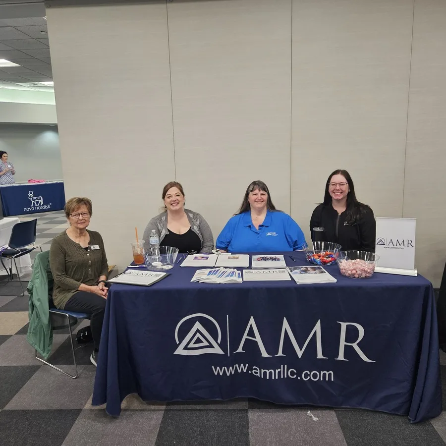 Four women sitting behind an AMR display table with promotional materials. They are smiling for the camera inside a large indoor venue.