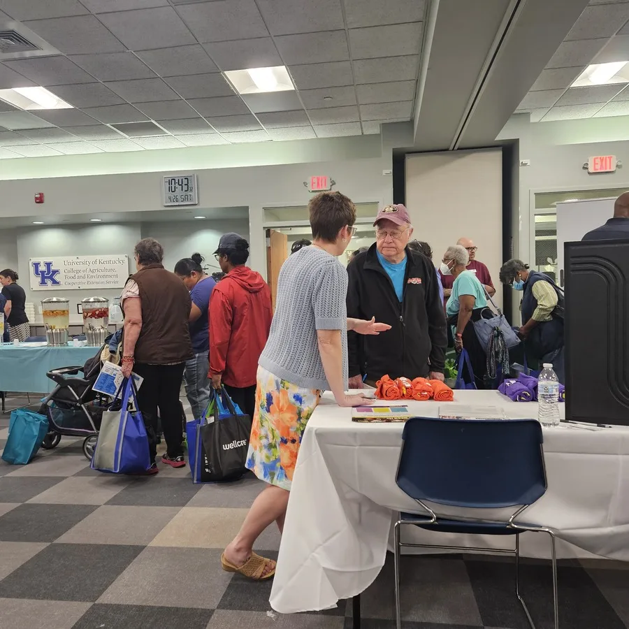 Crowded event space at the University of Kentucky with multiple attendees walking around. A woman in a floral skirt is talking to an elderly man at a booth.