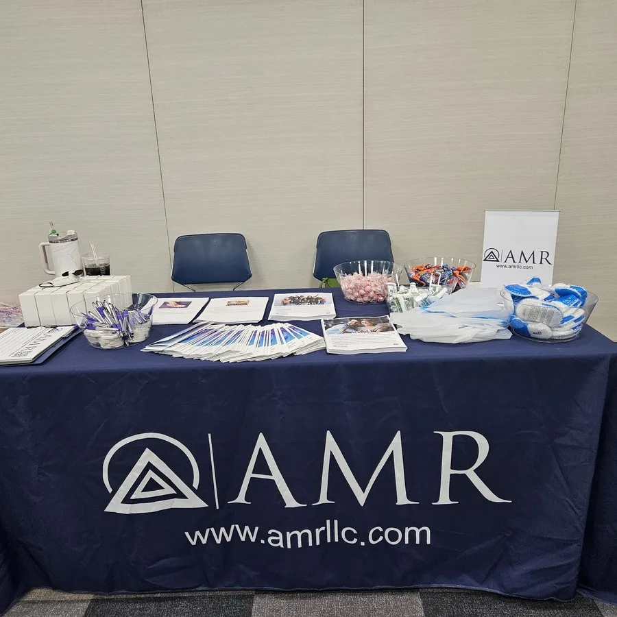 AMR booth table with two empty chairs, promotional handouts, pens, candy, socks, and branded signage, ready for attendees at an indoor event.