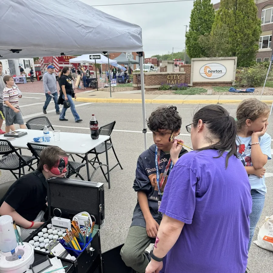 A face painting booth set up on a street during a community event outside Newton City Hall. A woman in a purple shirt is painting a young boy’s face while another child waits nearby. An artist with a red and black painted face sits beside a case full of brushes and paints. Folding tables and chairs are arranged under a canopy, with people and booths visible in the background along the street.