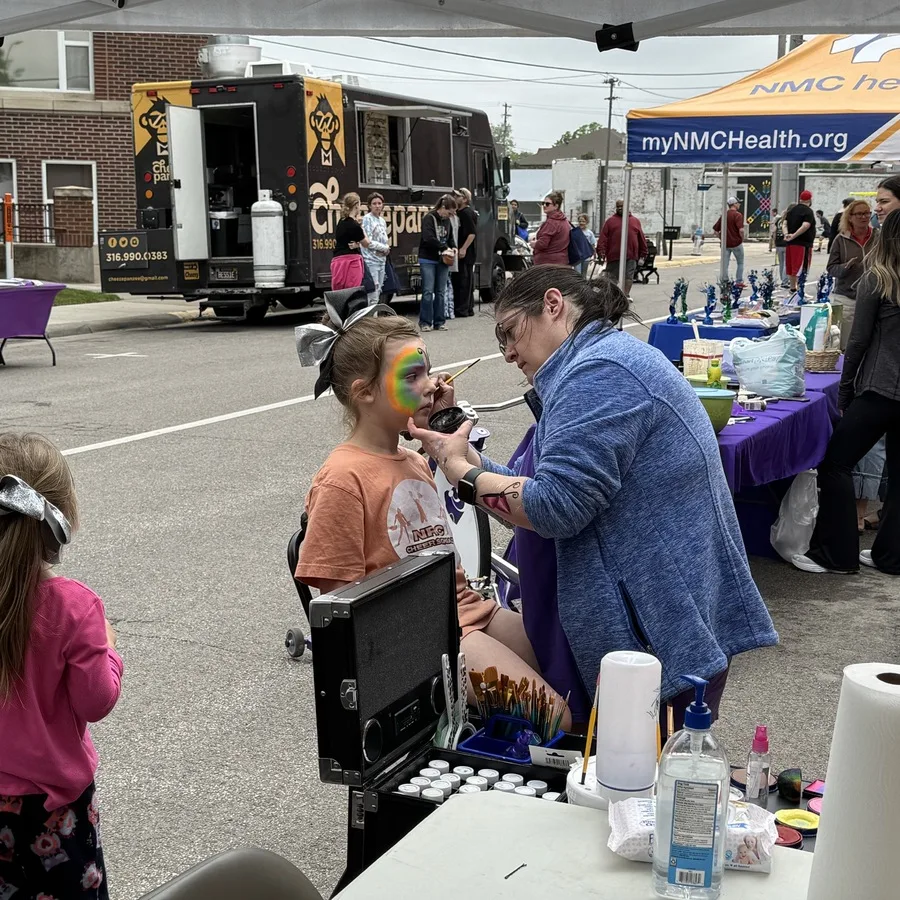 A face painter in a blue jacket is applying vibrant rainbow-colored face paint to a young girl wearing an orange t-shirt and a large silver bow. The booth is part of a lively street fair with food trucks, vendor tables, and a crowd in the background. A younger child in pink stands watching nearby. The artist’s table is stocked with brushes, paint palettes, and supplies.