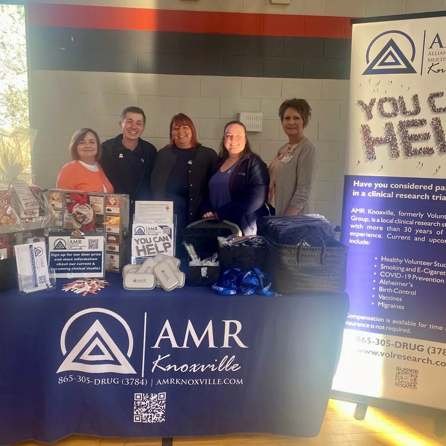 Six individuals are standing behind a professionally arranged AMR Knoxville booth indoors at a community expo. The table features a dark blue tablecloth with the AMR Knoxville logo, a QR code, and contact information. Promotional materials, brochures, gift baskets, branded giveaways, and a sign reading “You Can Help” are prominently displayed. A tall banner behind the table encourages participation in clinical research trials. The group is smiling under natural and overhead lighting.