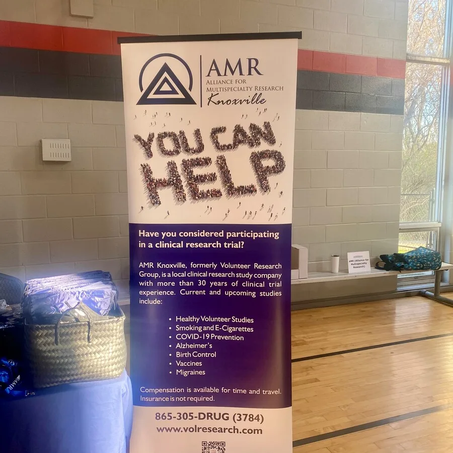 Close-up of a vertical banner from AMR Knoxville displayed at an indoor event. The banner features the AMR logo, the words “You Can Help” formed by a crowd of illustrated people, and details about clinical trial opportunities, including studies on COVID-19 prevention, smoking, Alzheimer’s, birth control, and more. Contact information and a QR code are included at the bottom.