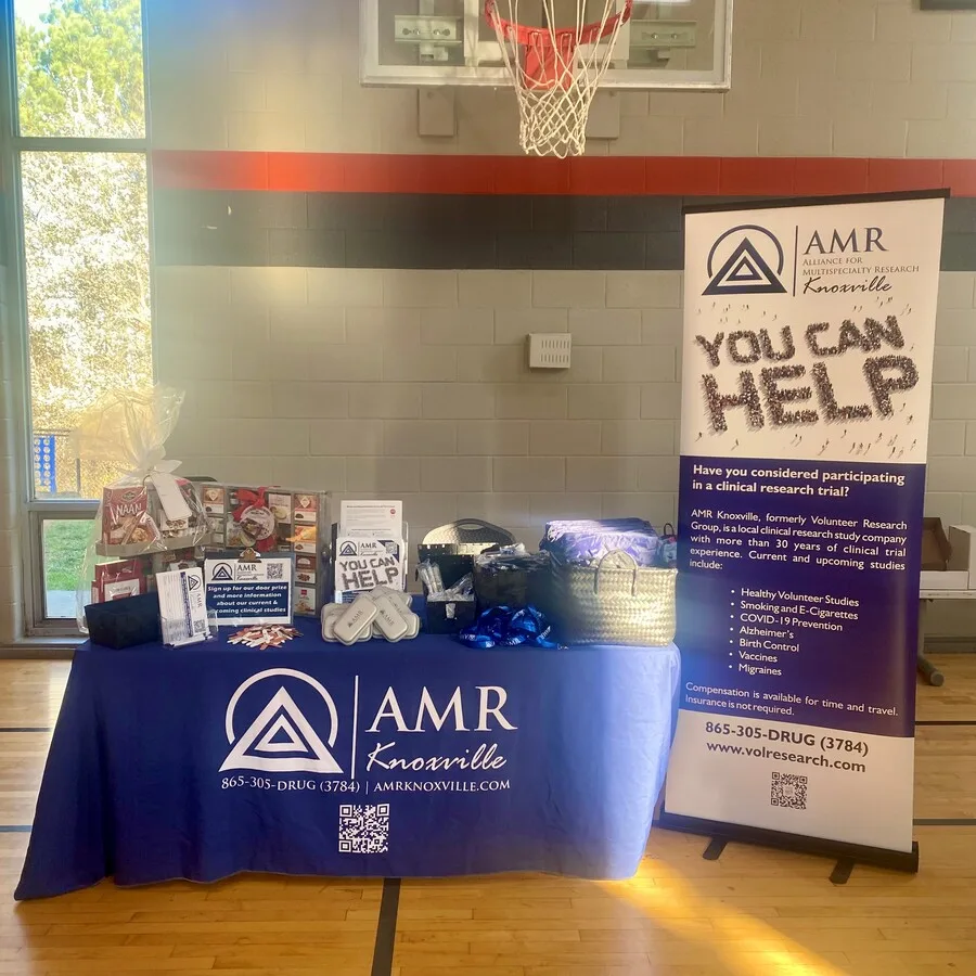 Wide shot of the AMR Knoxville booth set up inside a gymnasium beneath a basketball hoop. The table is covered in a navy tablecloth branded with the AMR Knoxville logo, QR code, and contact number. The setup includes a large gift basket, pamphlets, branded containers, and a sign-up sheet. A tall “You Can Help” banner stands next to the table, providing information about clinical trial participation.