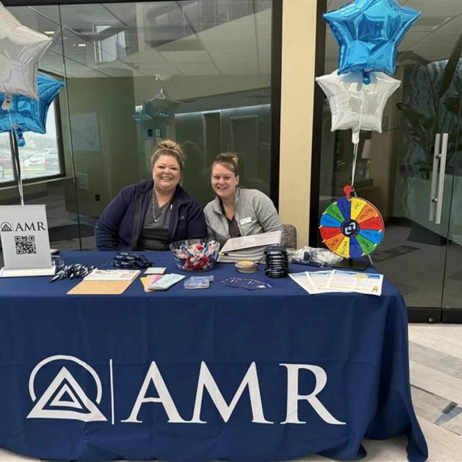 Two smiling women sit behind an AMR promotional table covered with a blue tablecloth bearing the AMR logo. The table is decorated with informational flyers, lanyards, pens, a bowl of candy, and branded items. Star-shaped blue and silver balloons are positioned at either side, and a colorful prize wheel is set up to the right. The setting appears to be an office or clinic lobby with glass doors and a modern interior.