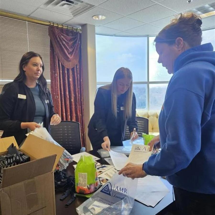 Three women assembling AMR event bags with flyers and promotional items in a room with large windows. One woman is handing a bag to another.