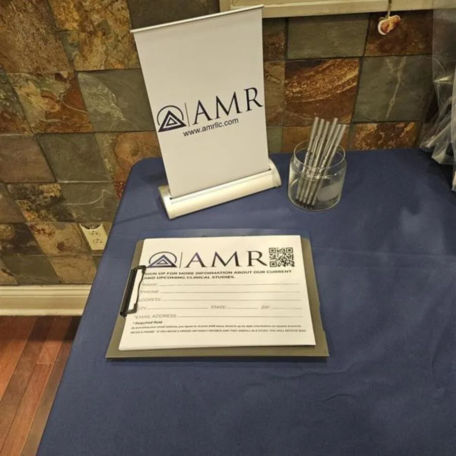Close-up of an AMR sign-up sheet on a clipboard beside a branded tabletop sign and a cup of pens, all on a blue-covered table.