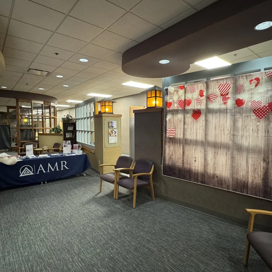 An AMR display table is set up in the corner of a cozy, carpeted waiting area with wooden chairs and warm lighting. In the background, a festive Valentine’s-themed photo backdrop is hung on the wall, decorated with red and pink fabric hearts against a wood-textured background. The table includes promotional materials and is partially visible on the left side of the image.