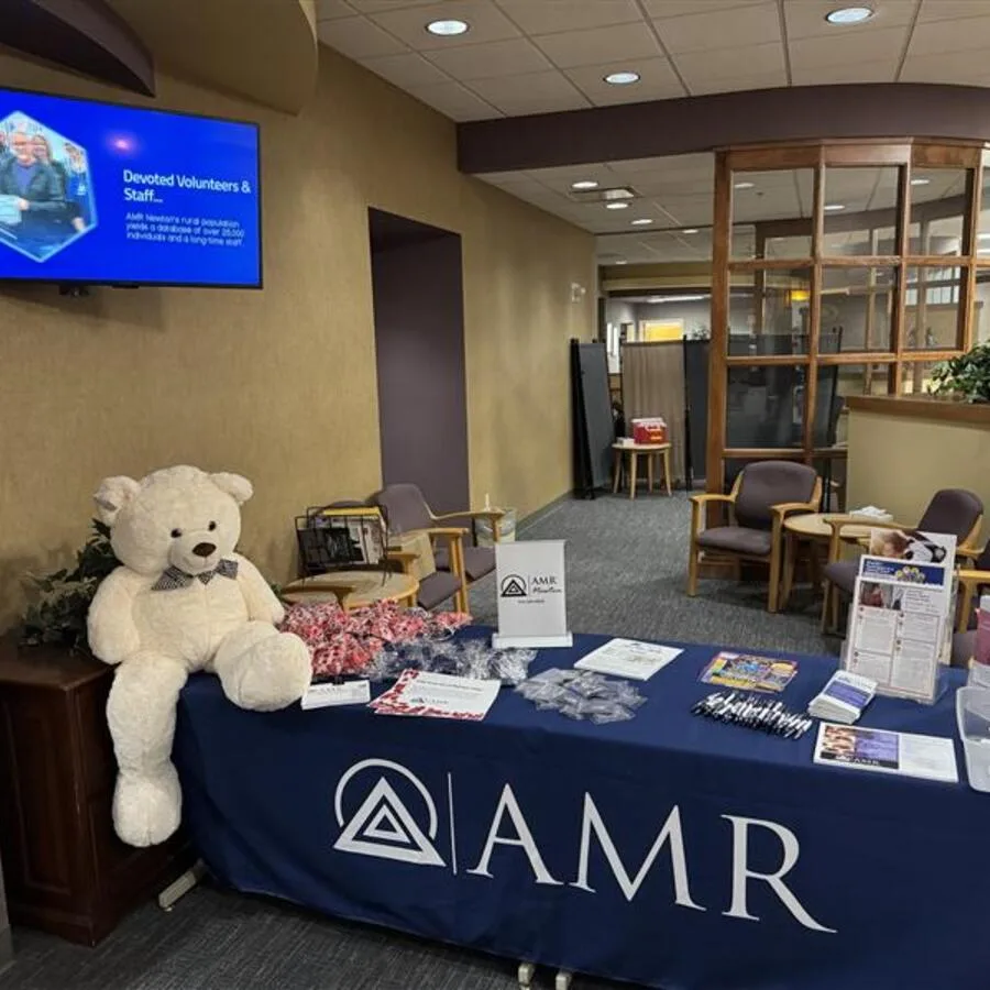 A large white teddy bear with a checkered bowtie sits beside an AMR display table covered with a dark blue tablecloth. The table is set up in a warmly lit lobby with informational brochures, candy, pens, and small giveaway items neatly arranged. A sign reading "AMR" and "Plains" is also visible on the table.