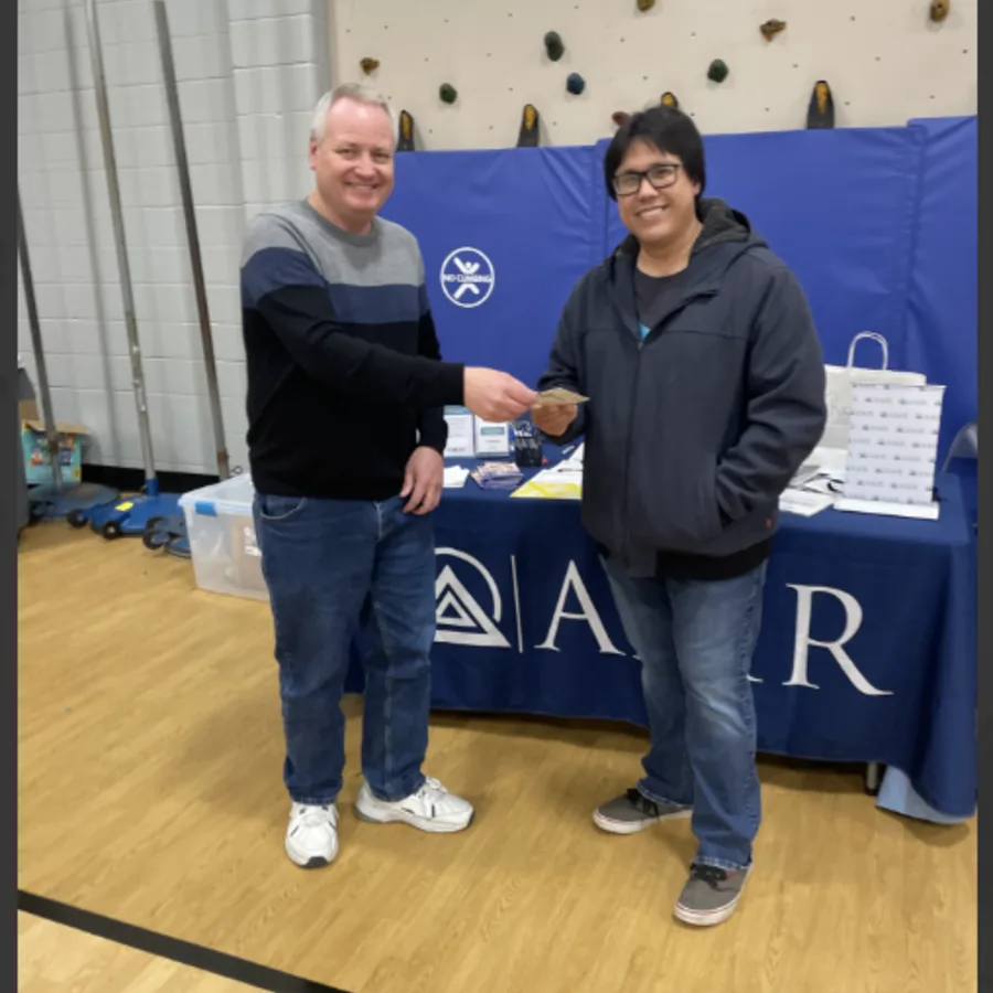 A man hands cash to another man in front of an AMR booth at an indoor event. They are both smiling, and the booth in the background features brochures and a white AMR gift bag. The floor is wooden, and a climbing wall is partially visible behind them.