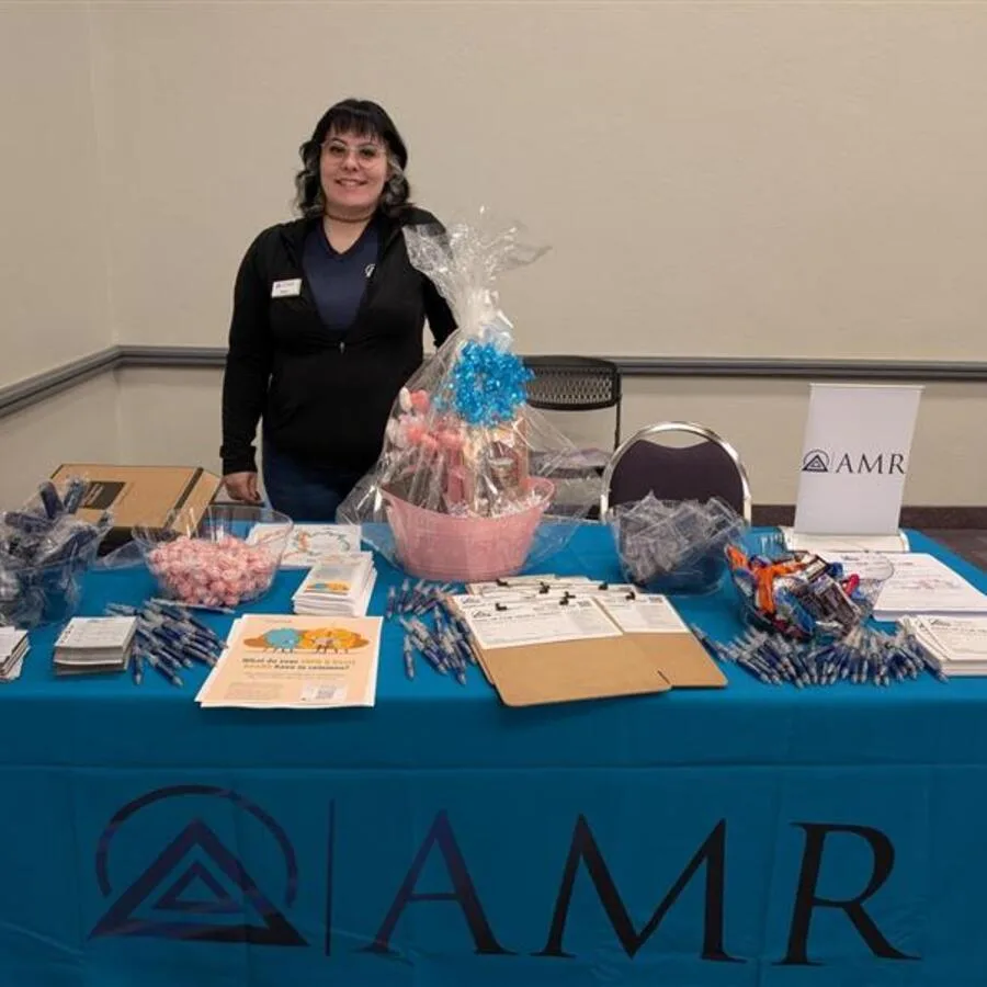 A woman stands smiling behind an AMR event booth indoors, wearing a black zip-up and name tag. The table is decorated with promotional materials such as pens, candy, forms, and a pink raffle basket with a blue bow. Blue and silver balloons are tied to the table, and branded signage is placed in the background.