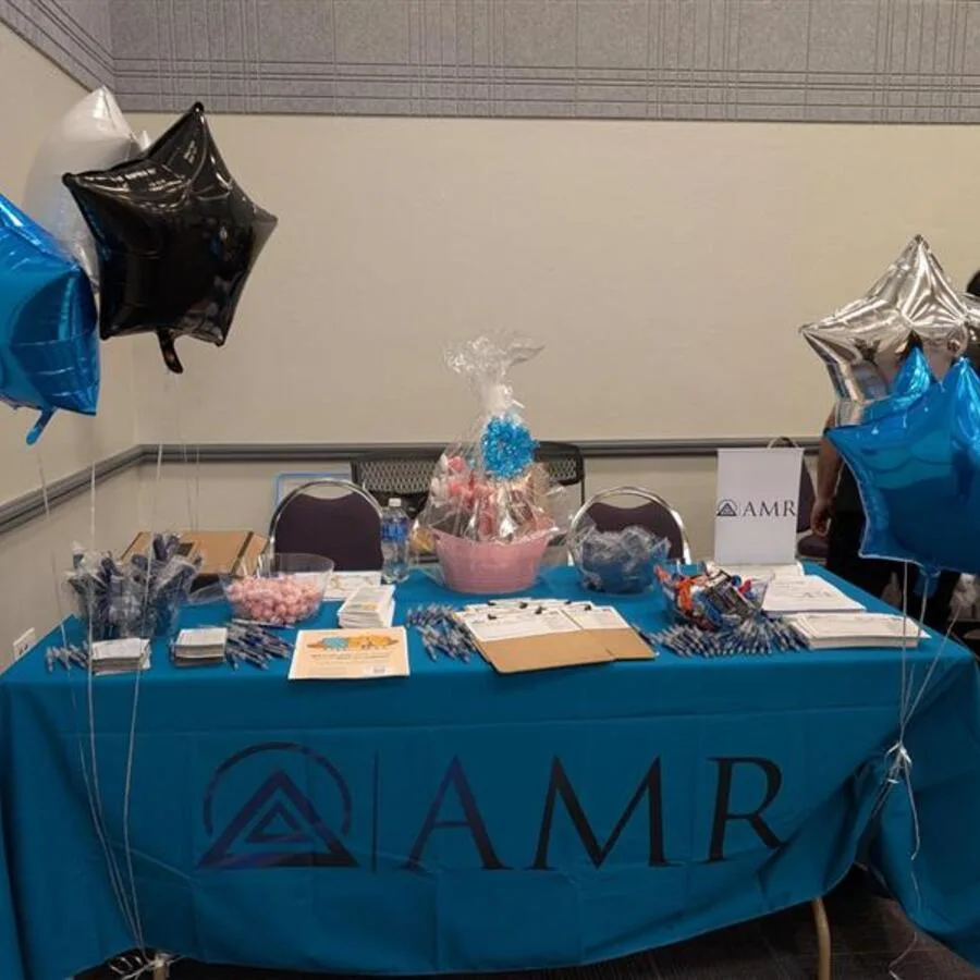 A blue AMR table covered with promotional items is displayed at an indoor event. The table is surrounded by metallic star-shaped balloons in black, blue, and silver. A large pink gift basket wrapped in plastic with a blue bow sits at the center. Pamphlets, pens, snacks, and registration forms are neatly arranged across the table, ready for attendees.