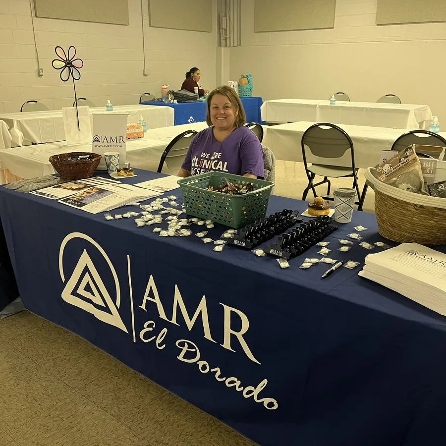 A woman wearing a purple “We Are Clinical Research” t-shirt sits behind an AMR El Dorado booth at an indoor community event. The table is covered with a navy blue tablecloth featuring the AMR logo and "El Dorado" in script. The booth is neatly arranged with promotional materials, small giveaway items like pens and mints, a basket of snacks, and informational brochures. Other tables covered in white and blue cloths are visible in the background, with one other person seated farther back.